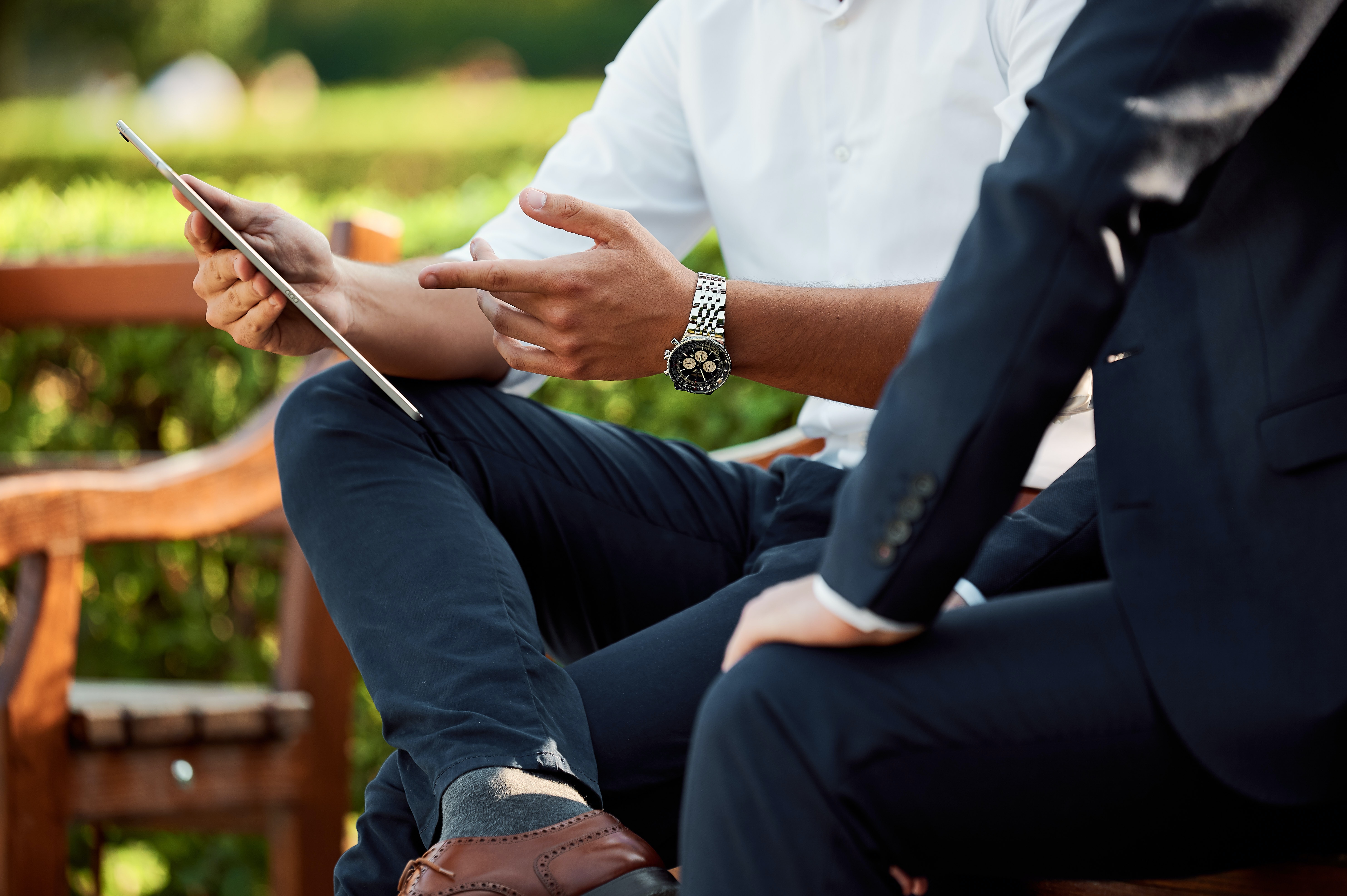 Two men sitting on a park bench ins summer on a bright sunny day.  Low bright green manicured low hedges in the background and a well-crafted wooden park bench to their right. The male on the left of the screen is wearing a bright white collared shirt with sleeves rolled up. Dark blue business causal pants blue socks and leather dress shoes. His right leg sits crossed over his left leg as he points to an image unseen on a tablet. The person to the right with hand on knee wearing a dark blue suit with four matching sleeve buttons appears to be listening attentively to the person in the white shirt. The image is cut off at their shoulders
